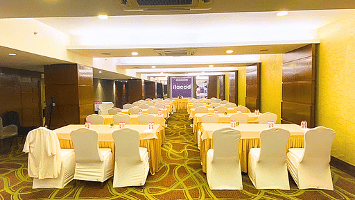 A conference room at Pride Plaza Ahmedabad, featuring rows of tables with white tablecloths, chairs, and a projector screen, suitable for meetings and presentations.